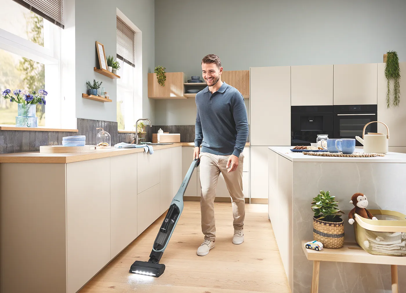A man in a modern European kitchen using the Bosch Flexxo Series 4 cordless stick vacuum (steel-blue color) on a wood floor, LED nozzle light visible