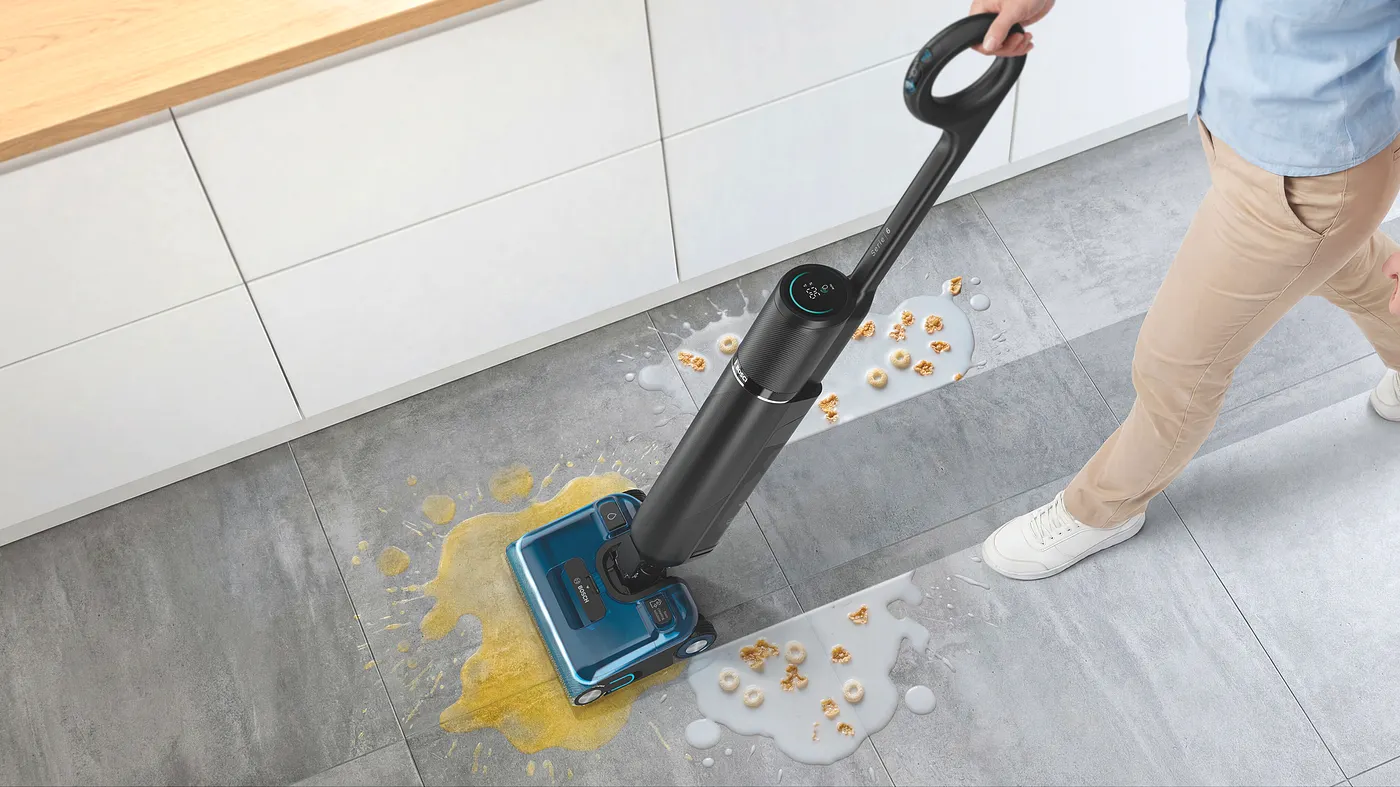 A person using the Bosch OneFlow Series 6 wet-and-dry vacuum to clean up a spilled bowl of cereal and milk on a grey-tile kitchen floor.