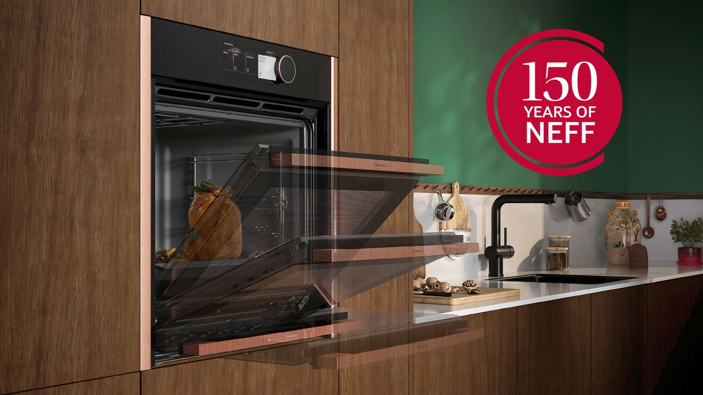 A NEFF built-in oven with its Slide & Hide door retracting beneath the oven in a wood-panelled kitchen, next to a red '150 Years of NEFF' badge