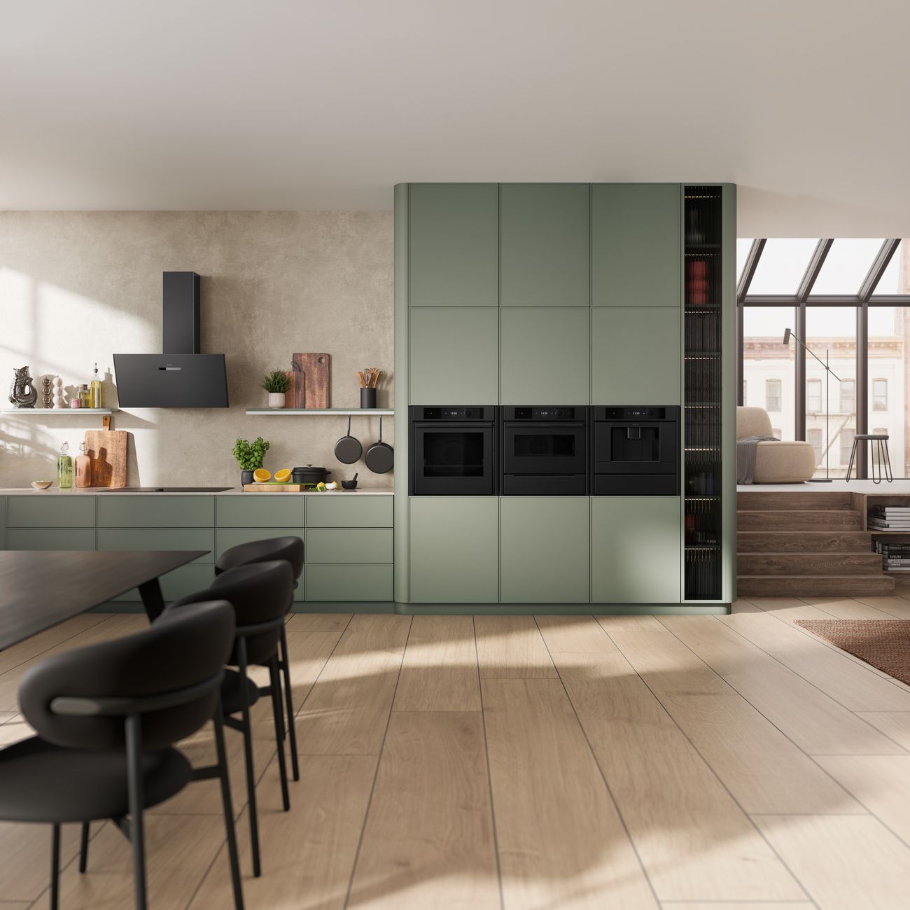 Open-plan kitchen with sage-green cabinetry, a matt black inclined hood on the wall, and a column of three matt black built-in appliances: an oven, a compact oven and a coffee machine.