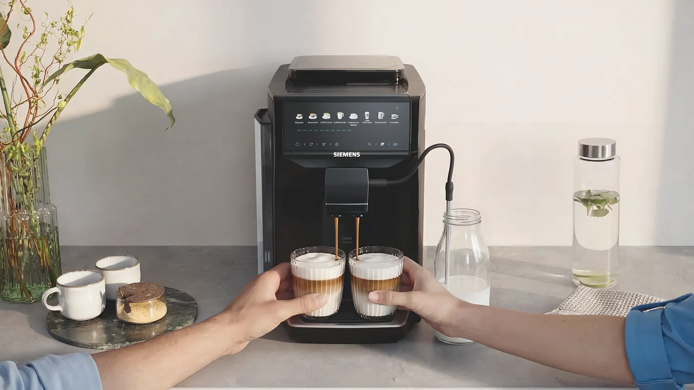Two hands taking two latte-macchiato-style coffee drinks from a Siemens EQ500 iAroma fully automatic coffee machine on a styled kitchen counter with plants and ceramic cups.