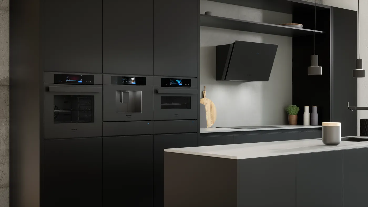 A modern matt-black European fitted kitchen showing a column of three built-in Siemens appliances in matt black finish, an oven, a coffee machine and a warming drawer, with a matching matt-black angled cooker hood above an induction hob and light countertop.